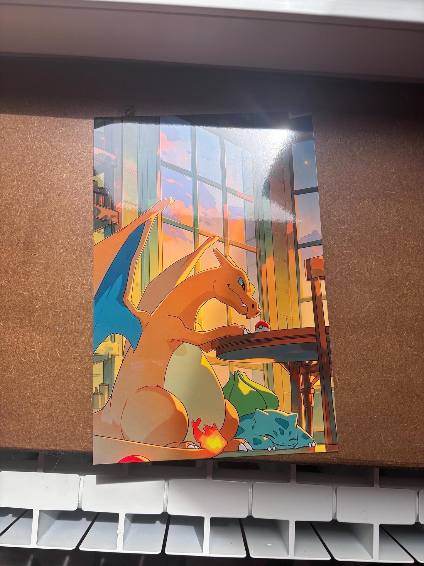 Peut inclure: Une impression d'art rectangulaire présentant une illustration numérique d'un Dracaufeu orange assis à une table en bois avec une petite Poké Ball rouge et blanche. Un Bulbizarre vert dort sur le sol sous la table dans une pièce ensoleillée.