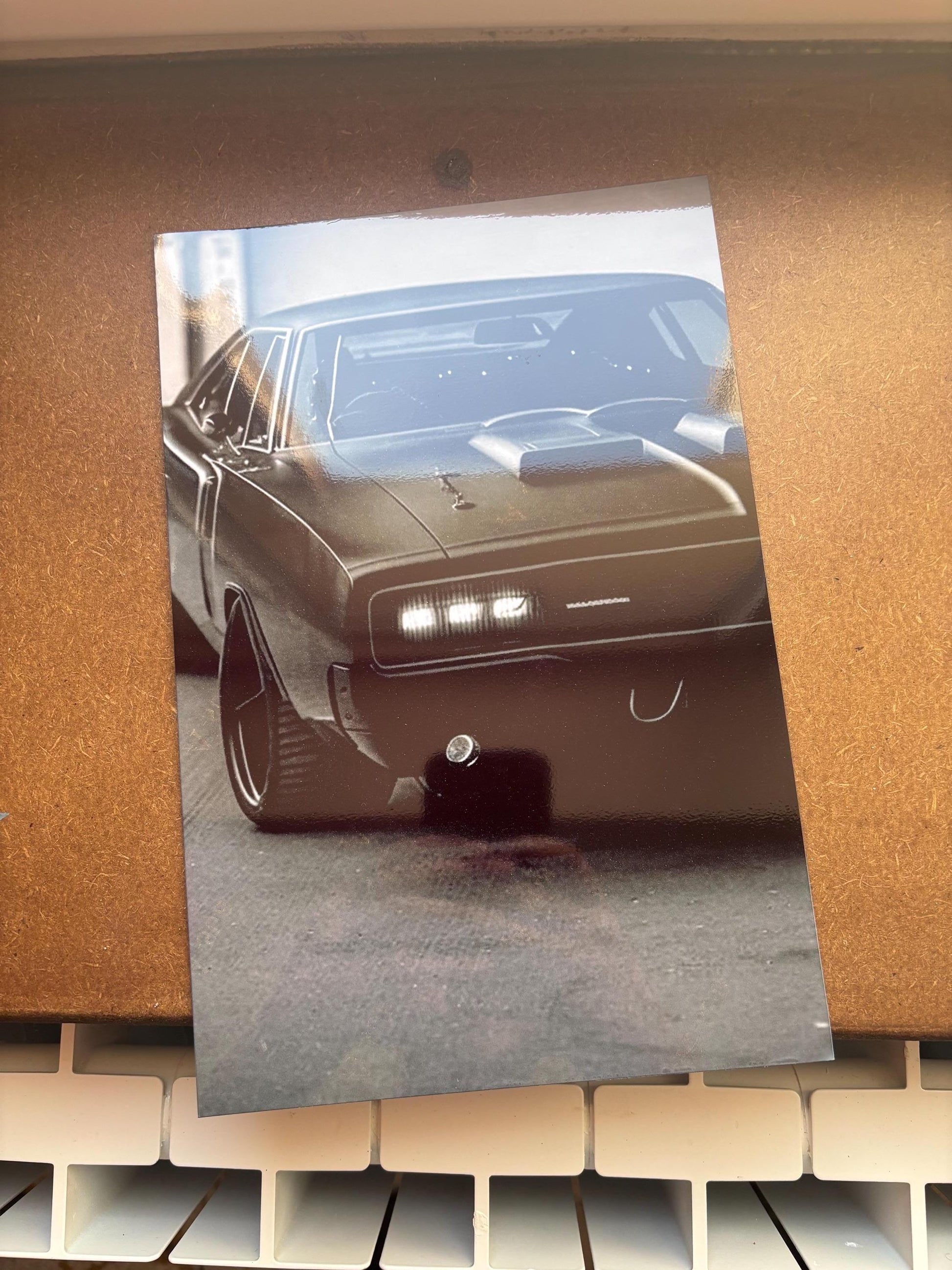 Peut inclure: Une impression rectangulaire brillante représentant une voiture de sport sombre vue de trois quarts avant. Le véhicule présente une prise d'air proéminente sur le capot, des phares rectangulaires à éclairage blanc et de grandes roues noires.